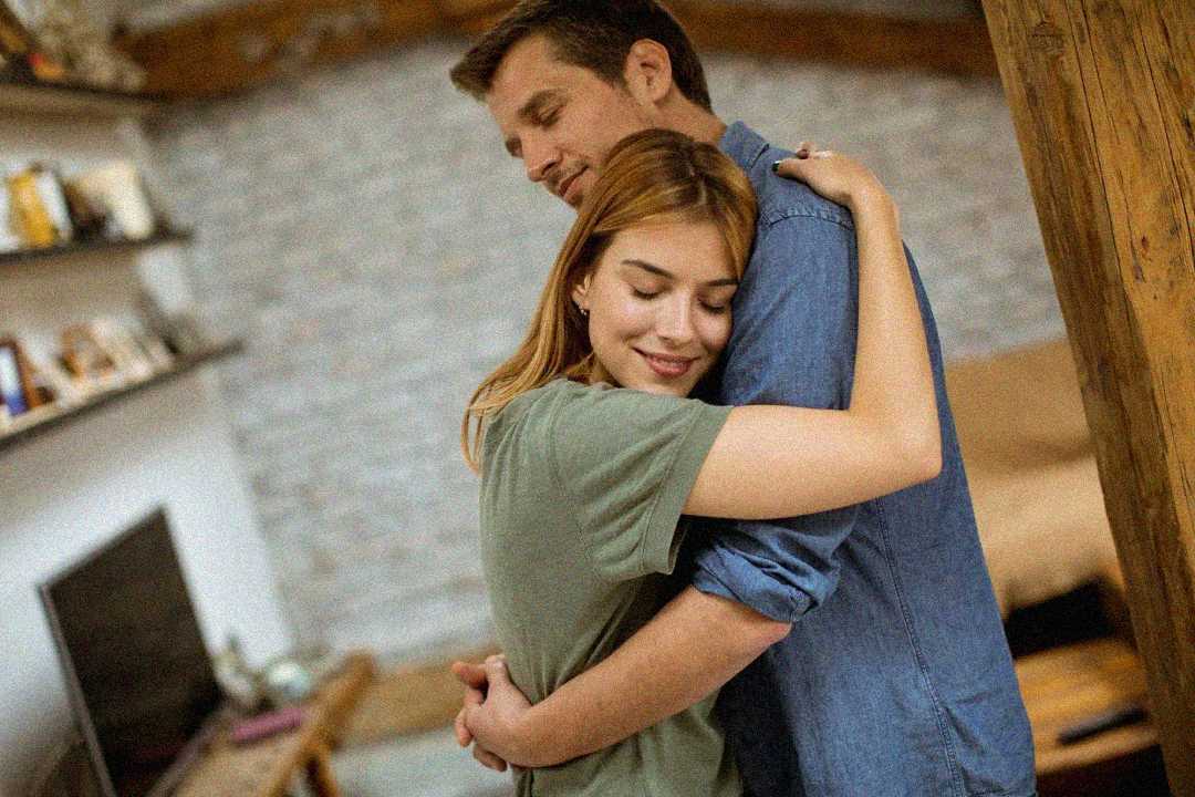 A young couple stands in a cozy living room, warmly embracing each other with eyes closed and peaceful expressions, conveying a sense of comfort and affection.