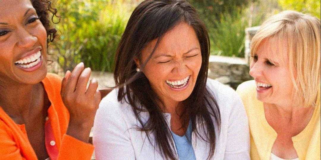 Three women sit close together outdoors, laughing and enjoying each other’s company. They wear colorful clothing and appear happy and relaxed in a sunlit setting.