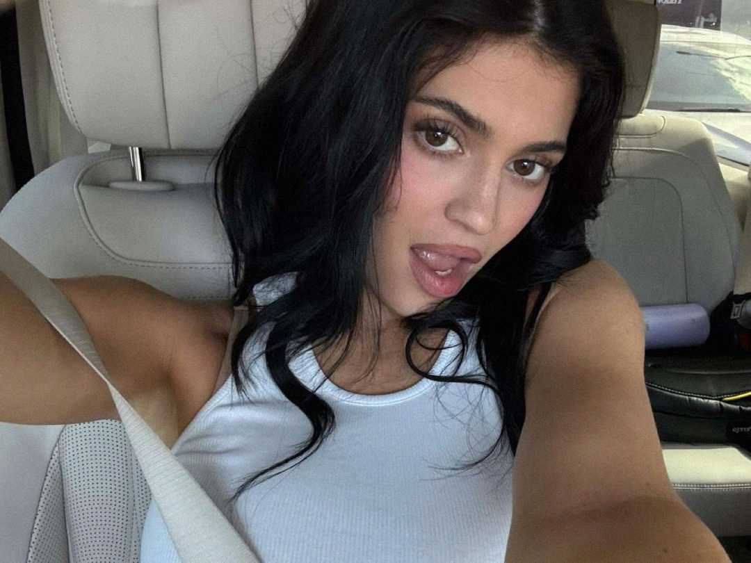 A woman with long dark hair wearing a white tank top takes a selfie in a car, looking at the camera with her mouth slightly open and her tongue out. She is wearing a seatbelt.