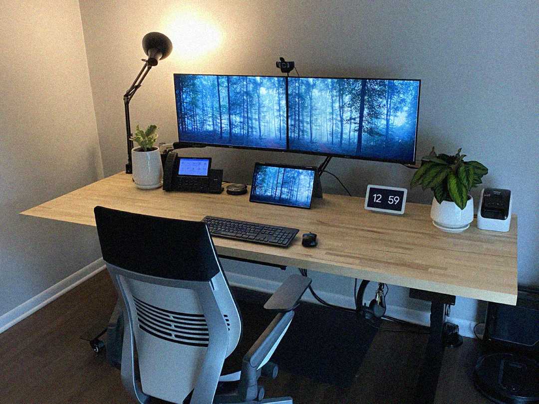 A modern desk setup with a wooden surface features two large monitors displaying a forest scene, a tablet, keyboard, mouse, desk lamp, clock, office chair, phone, and potted plants against a gray wall.
