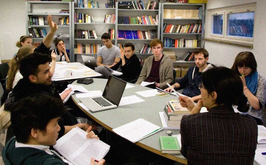 A group of students sit around a large oval table in a classroom, engaged in discussion with books, papers, and laptops. One student raises his hand. Bookshelves line the back wall.