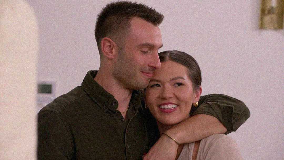 A man with short brown hair smiles, wrapping his arm around a woman with dark hair in a low bun. The woman is smiling, looking ahead, while the man looks down at her fondly. Both appear happy and close.