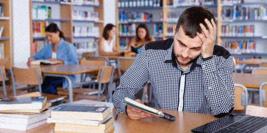A man sits at a desk in a library, looking stressed while holding a notebook. Several books and a smartphone are on the table, and other people are reading or studying in the background.
