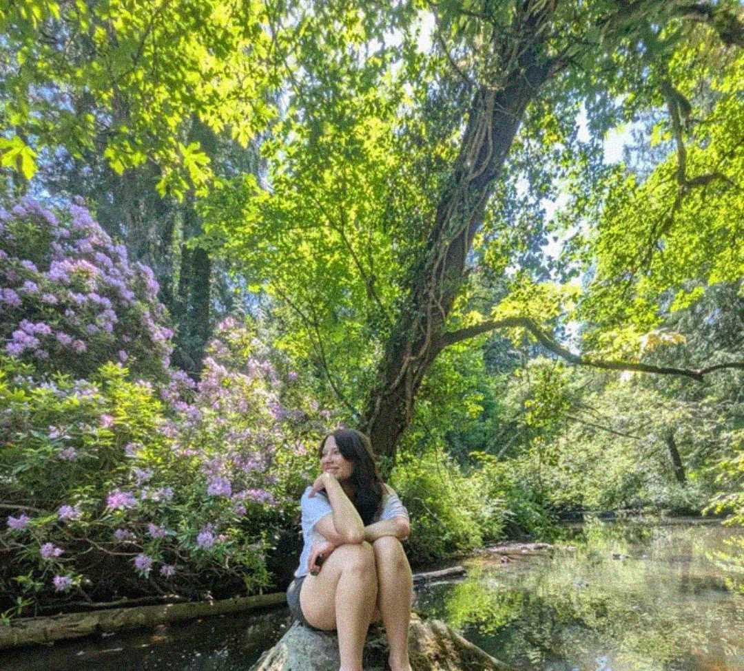 A woman sits on a rock beside a calm pond, surrounded by lush green trees and blooming purple flowers, with sunlight filtering through the foliage in a peaceful, natural setting.