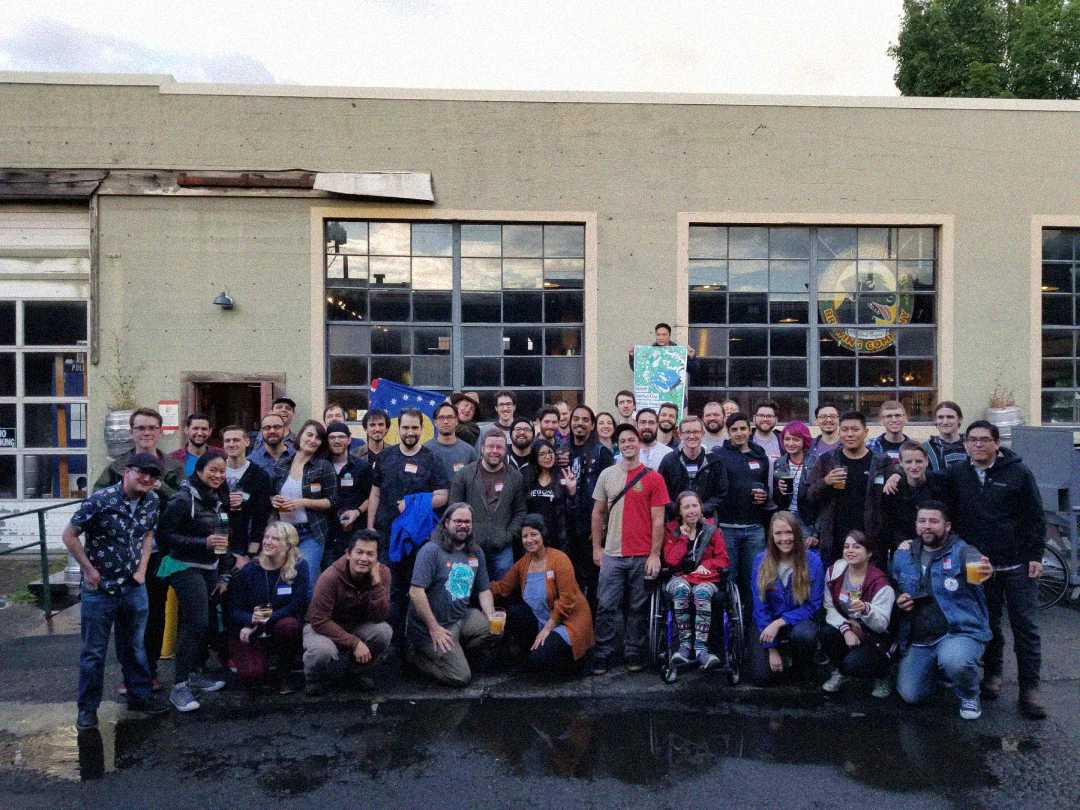 A large group of people pose together outside an industrial-style building with large windows, many holding drinks and smiling at the camera. The setting appears casual and festive, possibly a social or community event.