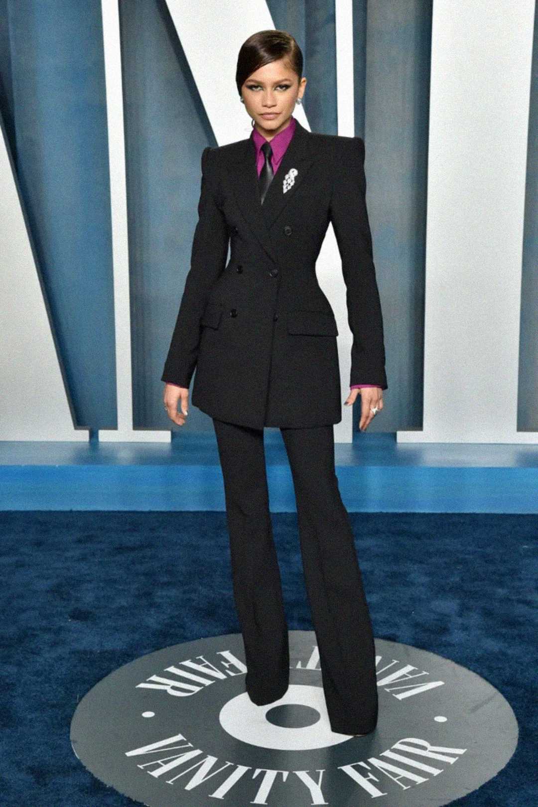 A woman stands on a blue carpet wearing a tailored black suit with flared pants, a magenta shirt, and a pocket square, posing confidently at a Vanity Fair event.