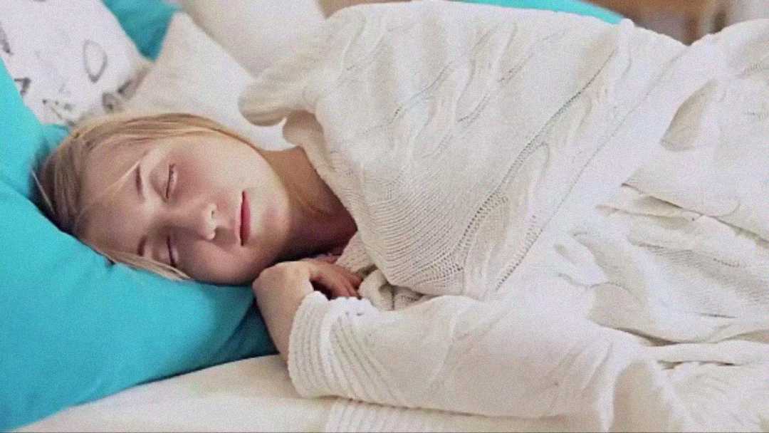 A person with light hair sleeps peacefully on a bed, covered with a white blanket and resting their head on a bright blue pillow.