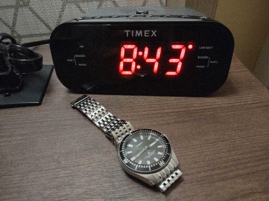A digital alarm clock displaying 8:43 in red numbers sits on a wooden surface next to a silver wristwatch with a black face and metal band.