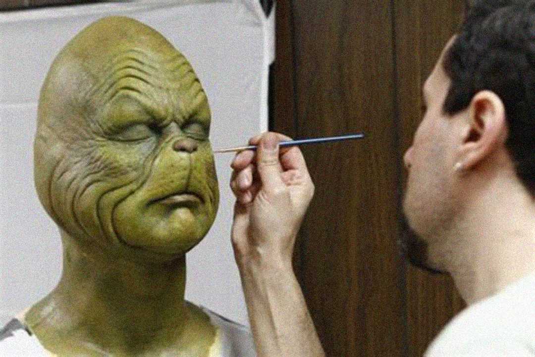 A person carefully paints details on a realistic green alien or creature mask worn by another person, likely for a film or costume, in a workshop or studio setting.