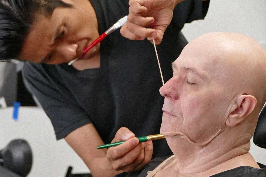 A makeup artist carefully applies a prosthetic or special effects makeup to a bald man's face, using tools and a brush, as the man sits with his eyes closed.