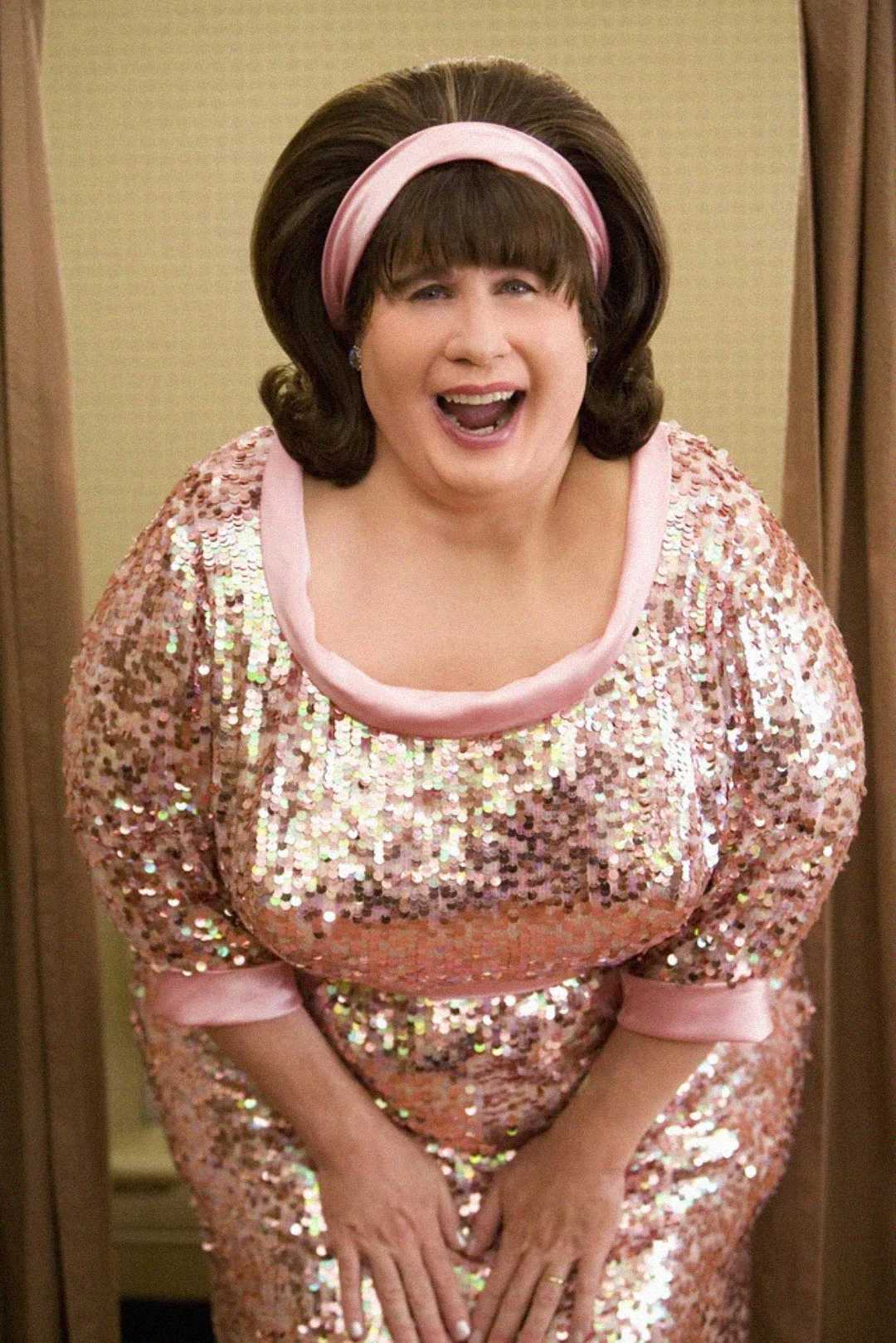 A person wearing a shiny pink sequin dress and a matching pink headband smiles widely, leaning slightly forward. They have short brown hair styled in a 1960s fashion. The background is neutral with beige curtains.
