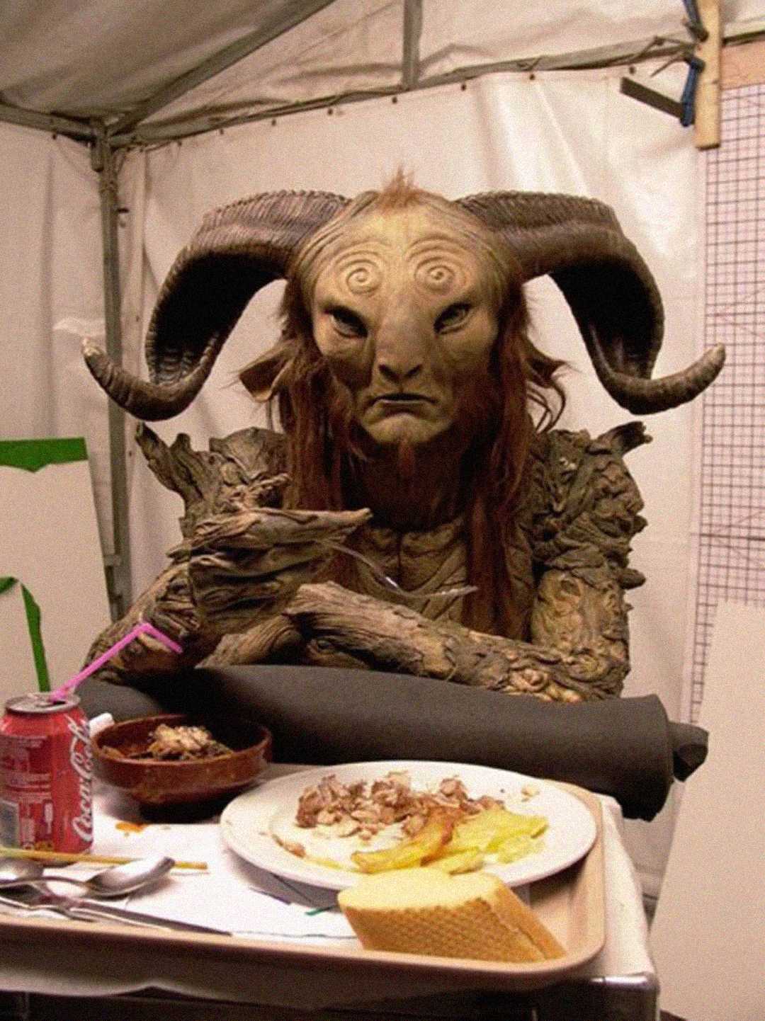 A person in an elaborate faun costume with large, twisted horns sits at a table in a tent, eating a meal with a can of Coca-Cola and dishes of food in front of them.