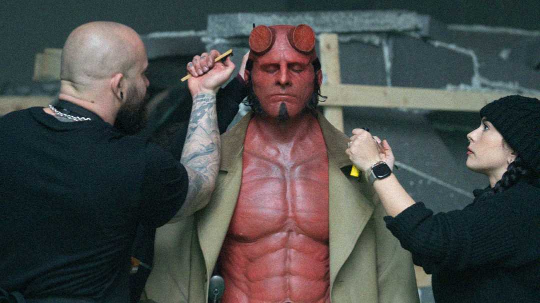 A man in red body paint and prosthetic horns, dressed as Hellboy, stands still with his eyes closed while two makeup artists touch up his makeup and costume on a film set.