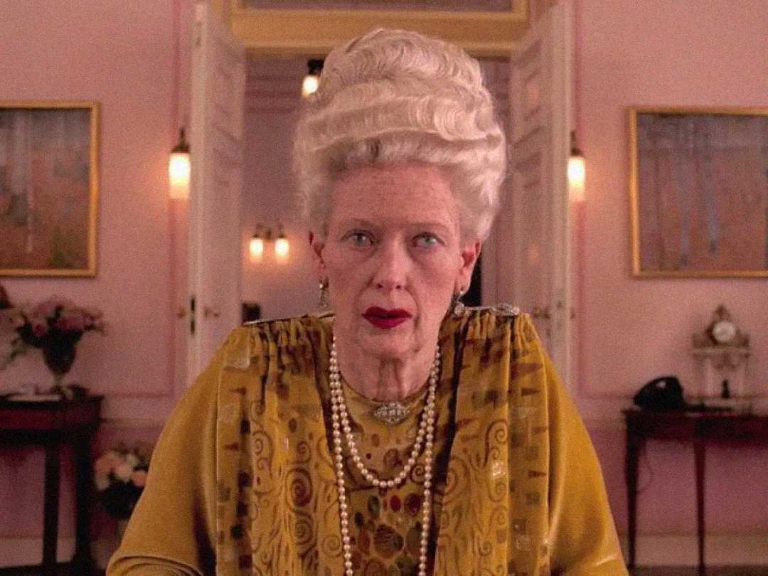 An elderly woman with white hair styled in an elaborate updo sits in a pink room. She wears a gold patterned dress, layered necklaces, and red lipstick, looking directly at the camera with a serious expression.