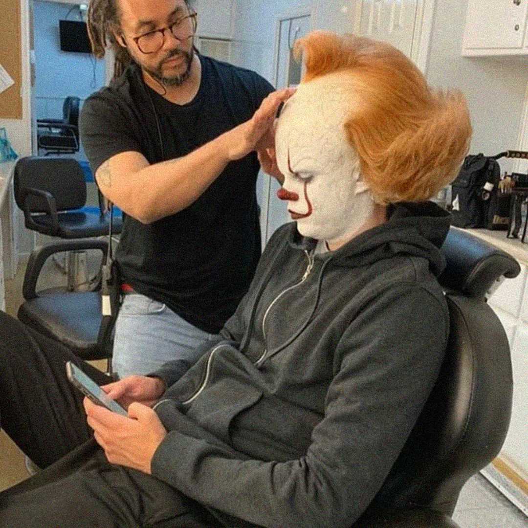 A makeup artist adjusts the wig of a person dressed as Pennywise the clown from "It," who is sitting in a chair and looking at a phone in a makeup room.