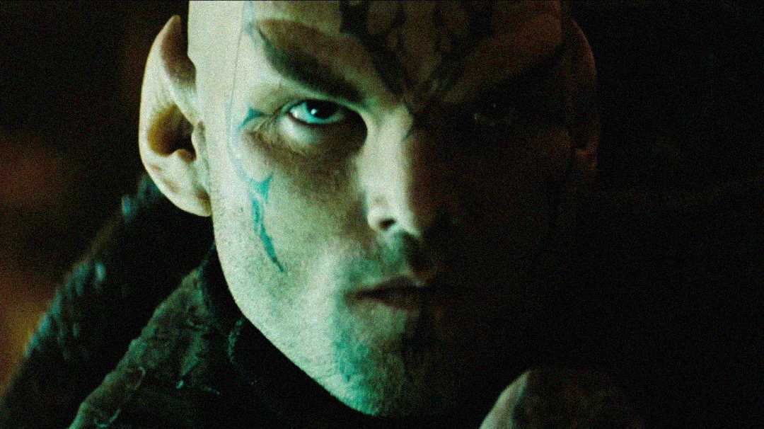A close-up of a person with a stern expression, prominent facial tattoos, pointed ears, and a shaved head, wearing a dark, textured outfit in dim lighting.