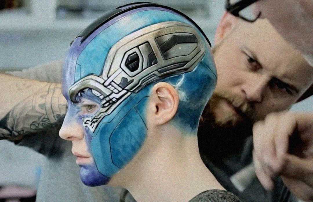 A makeup artist works on a person with their head and face painted in blue and silver, resembling a robotic character. The artist is focused, holding a tool near the person's head.