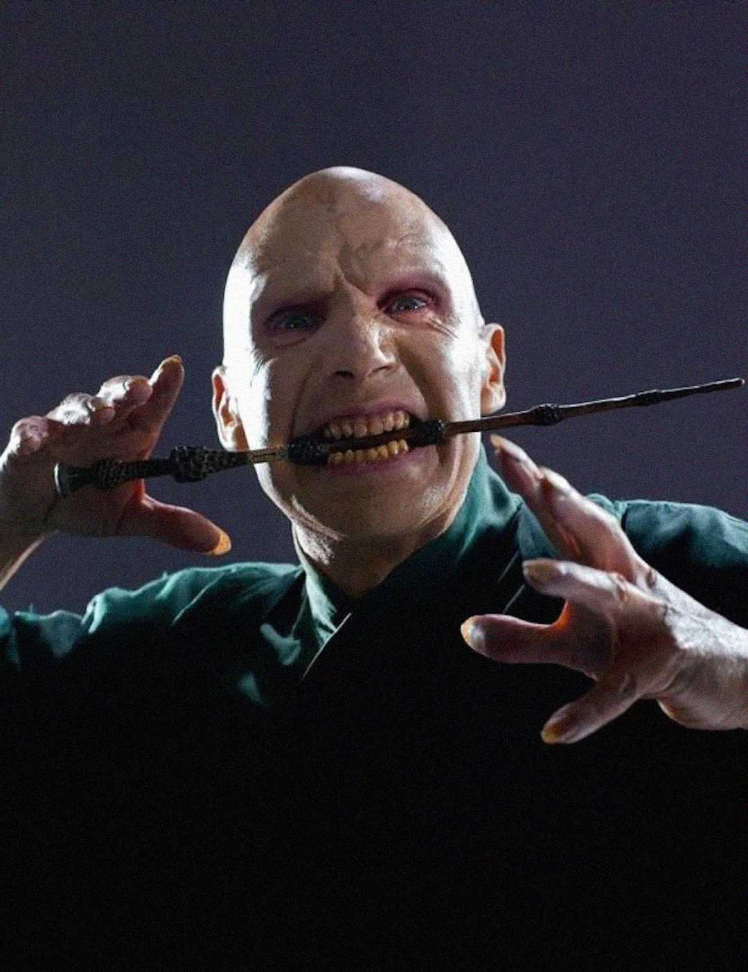 A bald, pale-skinned person with red eyes holds a wand between their teeth while making a claw-like gesture with one hand, set against a dark background.