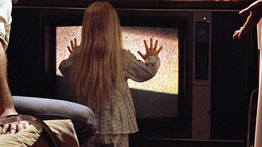 A young girl in pajamas stands with her hands pressed against a television screen showing static, while an adult sits nearby and another person’s arm is visible to the right.