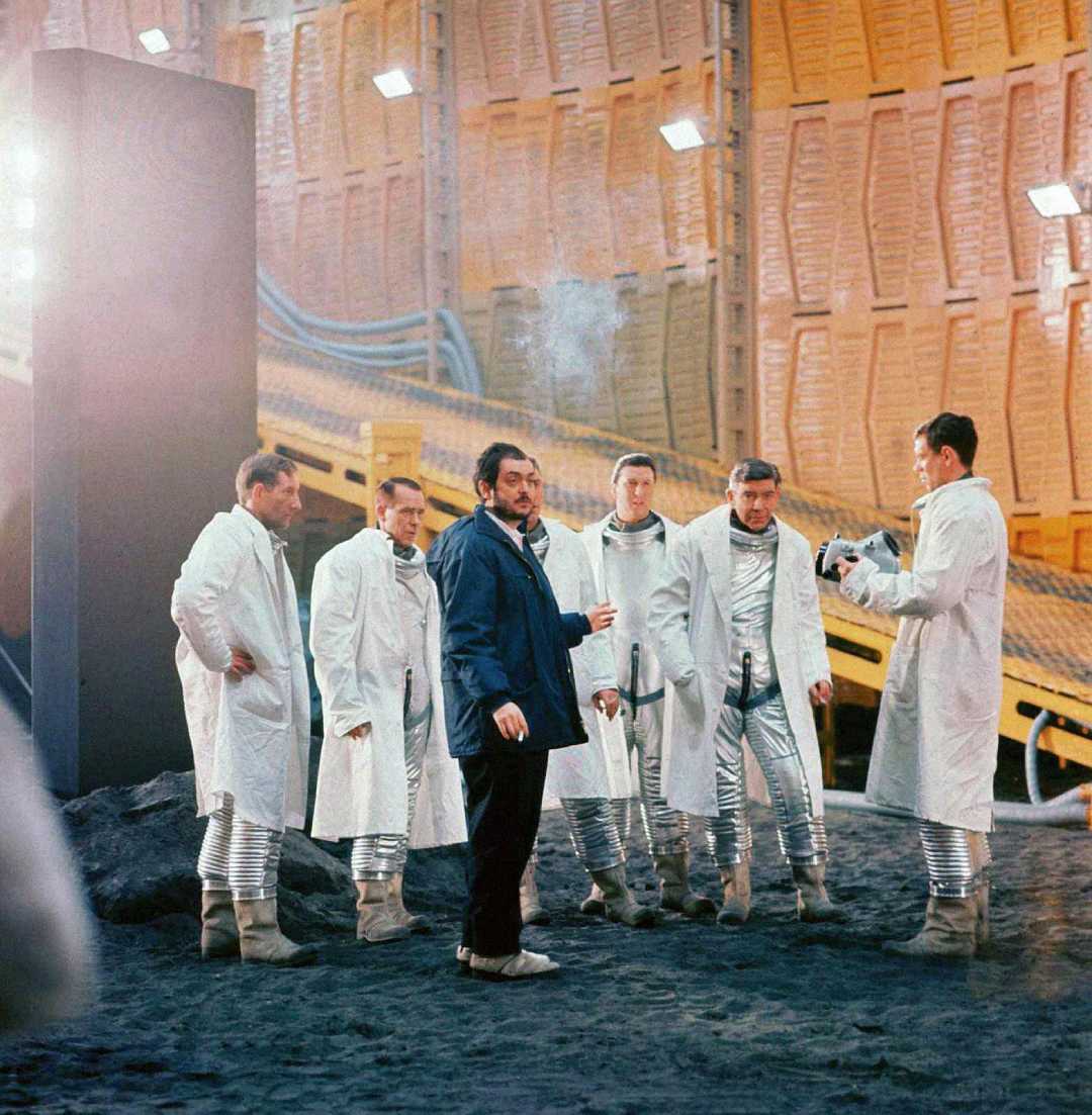 A group of men, some in futuristic silver suits and others in white coats, stand on a dark set with industrial orange walls. A man in the center, holding a cigarette, appears to give instructions. Bright light shines from the left.