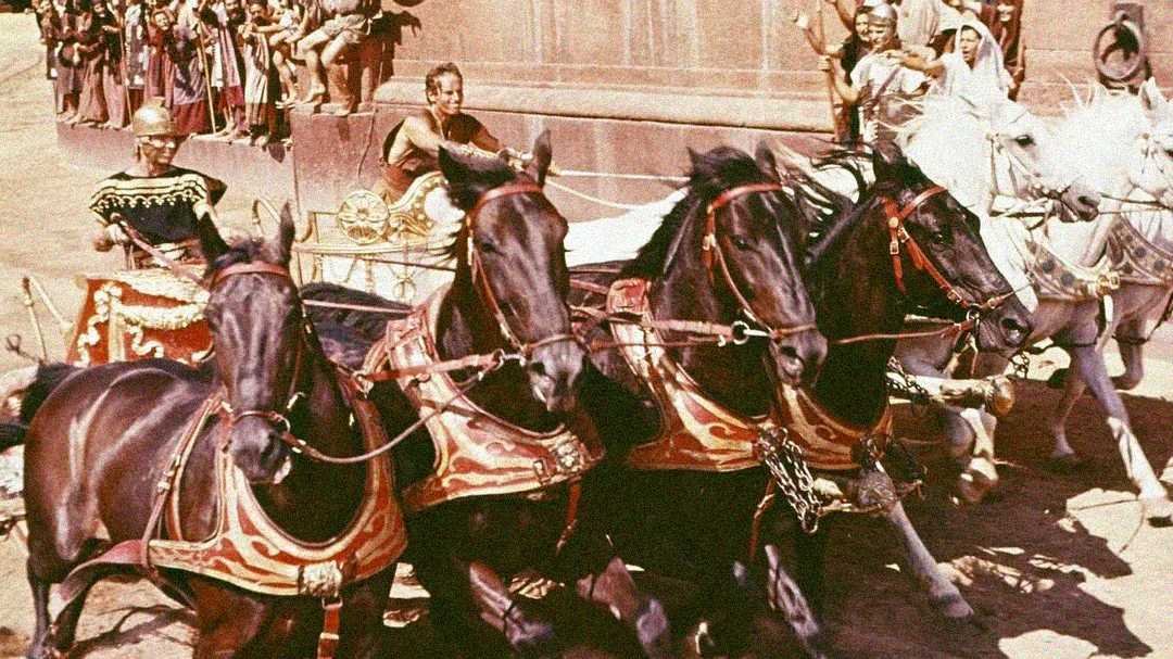 A dramatic chariot race scene with four black horses pulling a chariot, driven by a man in ancient attire, with another chariot and spectators in the background.