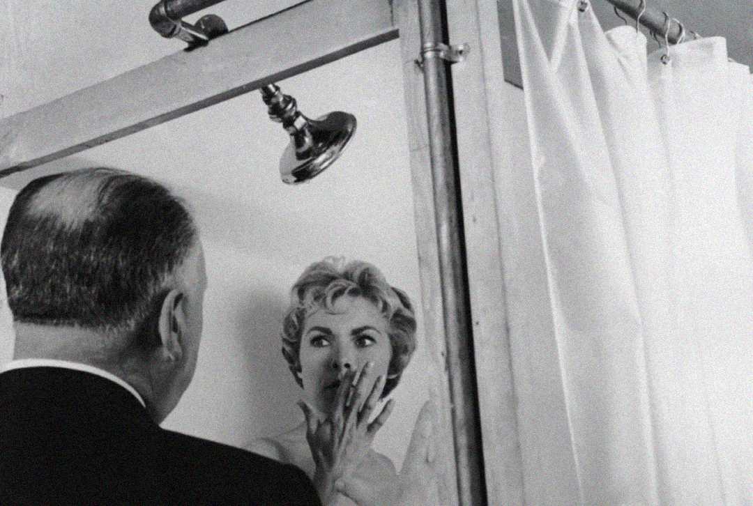 A black-and-white photo shows a man with his back to the camera standing in a bathroom set, speaking to a startled woman behind a shower curtain, her hand covering her mouth. A showerhead is visible above them.