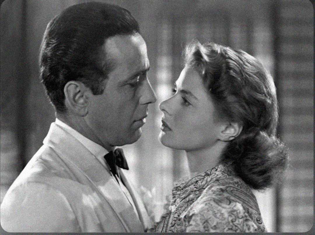 A black-and-white photo of a man in a suit and bow tie gazing intently at a woman with wavy hair, who is looking back at him closely. They appear serious and are standing very near each other.