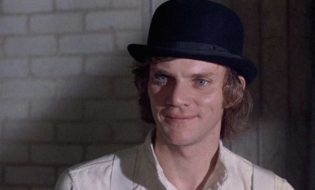 A young man with blue eyes wears a black bowler hat and a white shirt, smiling slightly while standing in front of a pale brick wall.