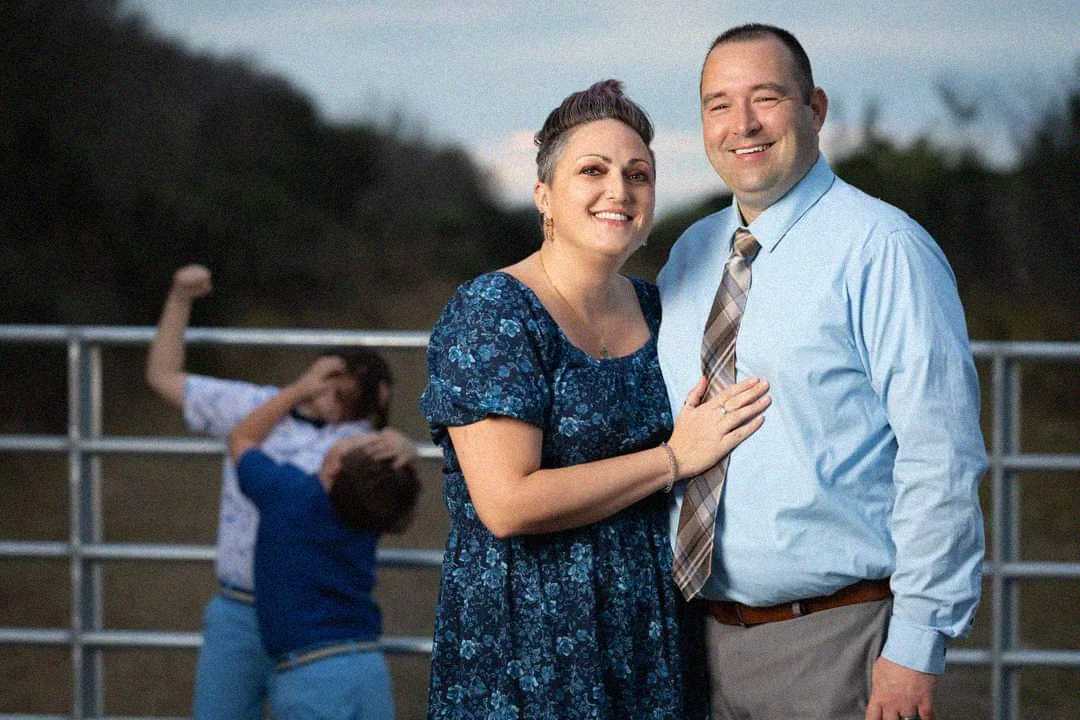 A smiling couple poses in the foreground, while two children play-fight in the blurry background near a metal fence outdoors at dusk.
