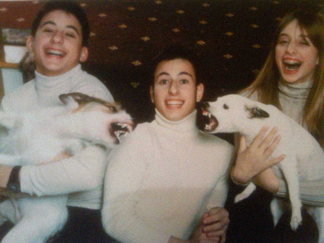 Three people in matching light turtlenecks sit together, smiling and laughing, while holding two dogs that appear to be playfully barking or growling at each other. The background shows a patterned wall.
