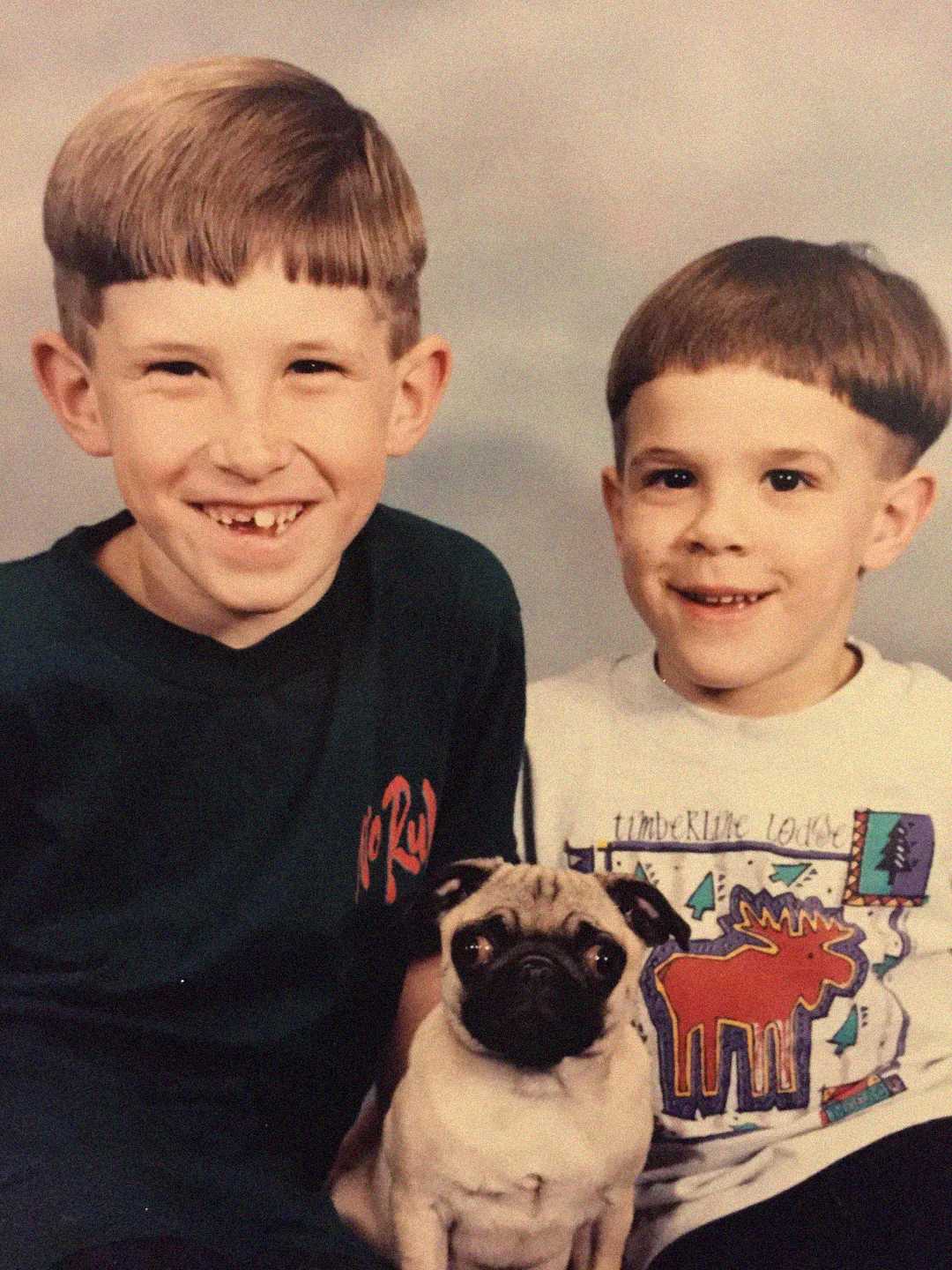 Two young boys with bowl haircuts, one wearing a dark shirt and the other a graphic T-shirt, smile at the camera while a small pug dog sits in front of them, all posing for a studio portrait.