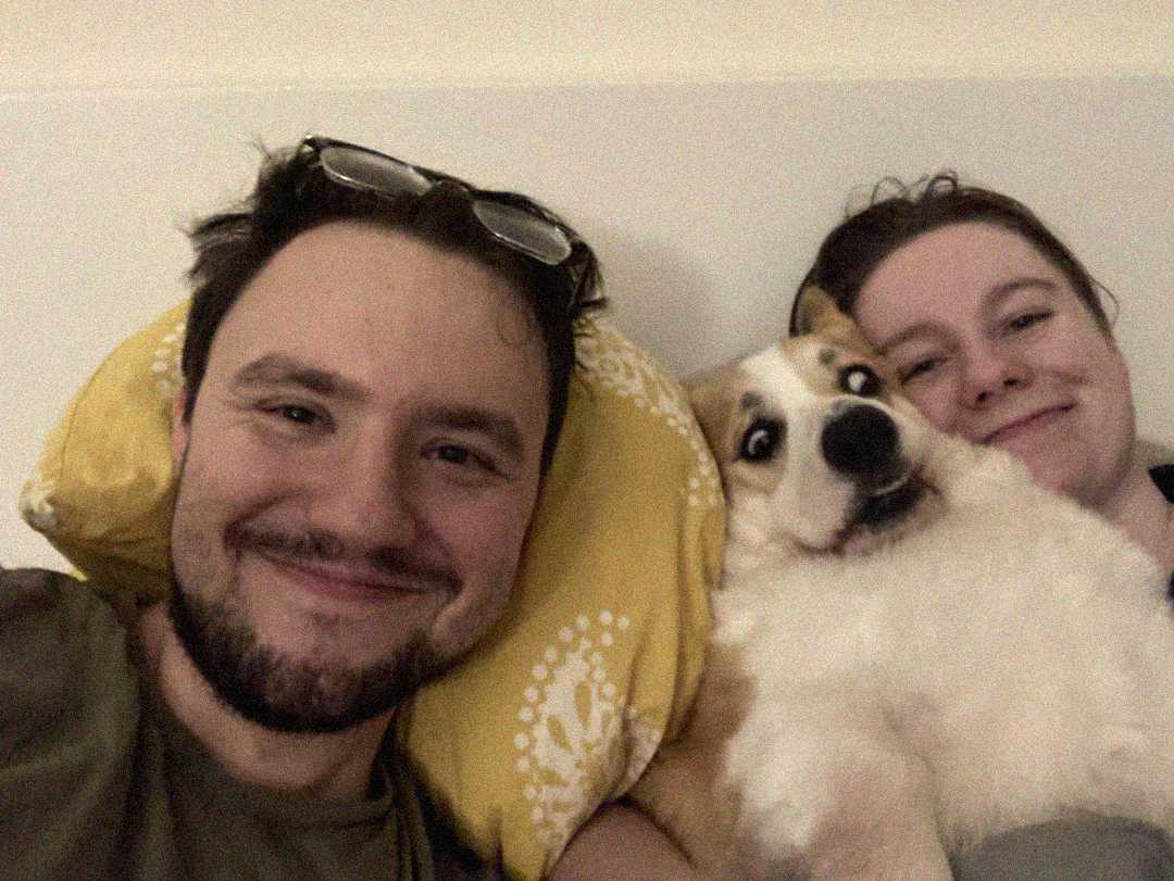 A man and woman are lying on a bed, smiling at the camera, with a fluffy dog between them. The man has glasses on his head and the dog looks wide-eyed. The woman rests her head on a yellow pillow with a white pattern.