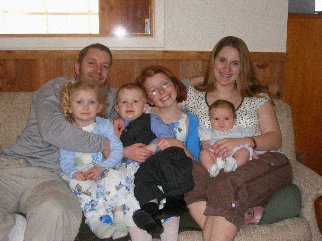 A family of six sits on a couch, smiling. Two adults sit at each end, with three young children and a baby in between them. The setting appears to be a cozy living room with wood paneling.