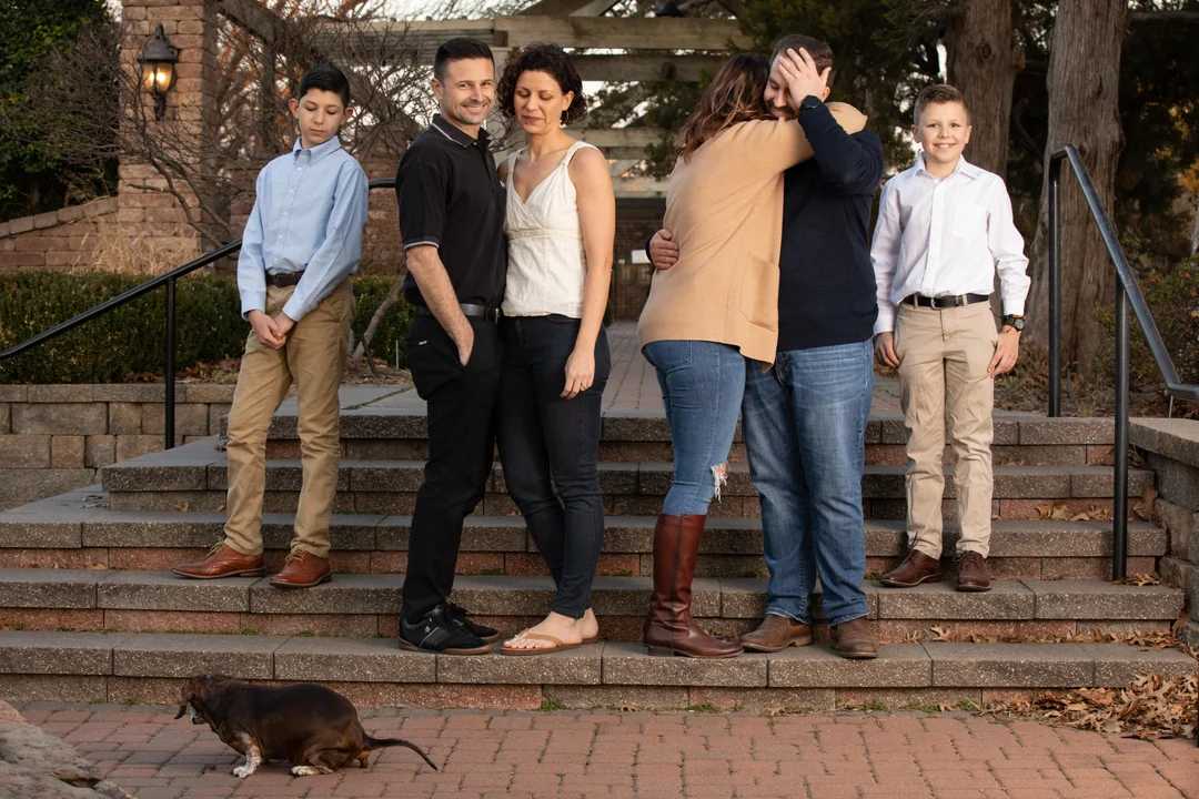 Six people pose on outdoor stone steps; two couples stand close, one pair hugging, while two boys stand apart. A small brown dachshund sniffs the ground in the foreground. Trees and stone walls are in the background.