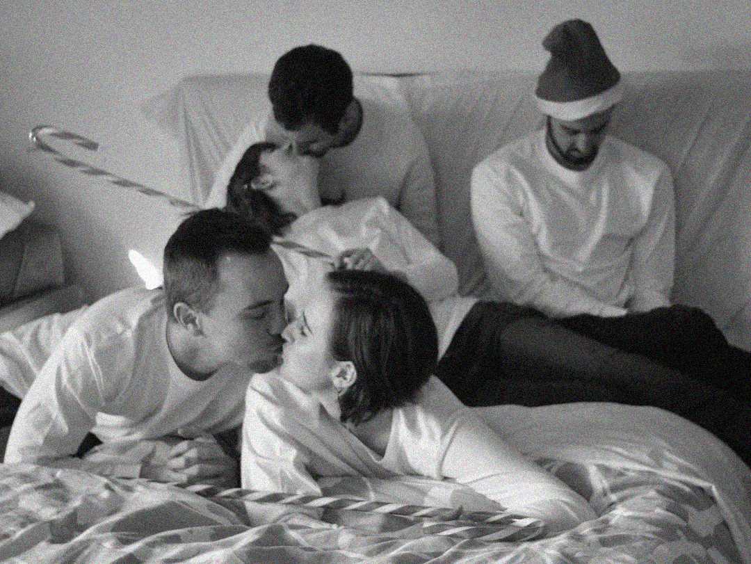 Five people sit on a couch and bed; two couples in white shirts kiss, while the fifth person, wearing a Santa hat, sits alone looking down sadly. A large candy cane prop is also visible. The photo is in black and white.