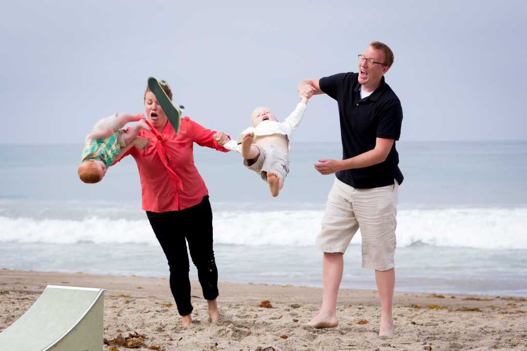 A woman and man on a beach accidentally let go of two small children, who are midair in an awkward moment; the ocean and cloudy sky are in the background.