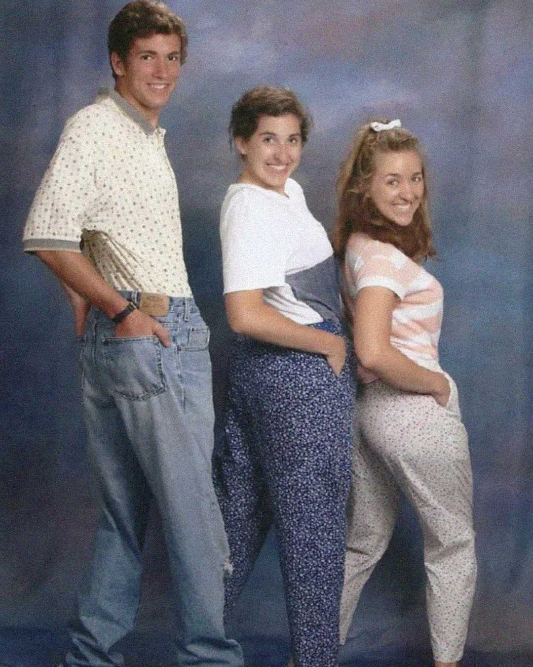 Three young people pose playfully in front of a studio backdrop, each with a hand in their pants pockets, smiling at the camera and dressed in 90s-style casual clothing.