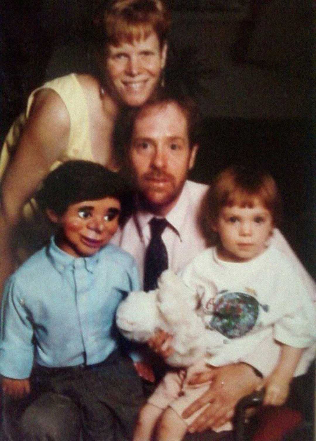 A woman stands behind a man who is seated with a young child holding a stuffed animal and a ventriloquist dummy, all posing together and smiling for a family portrait.
