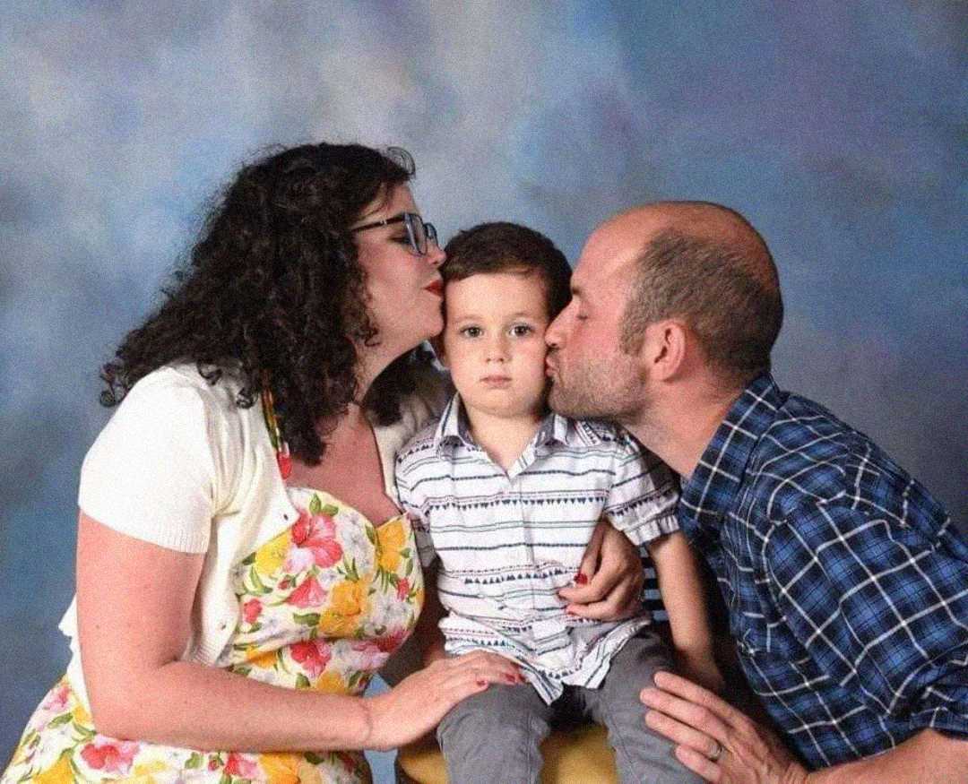 A woman and a man kiss a young boy on each cheek. The boy sits between them with a neutral expression, while the adults smile. They are in front of a blue and purple studio backdrop.