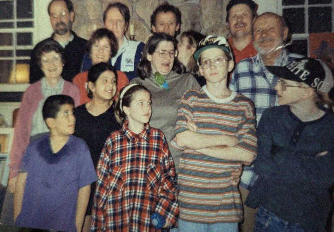 A group of people, including children and adults, are gathered indoors, posing closely together and smiling. Most are wearing casual clothes, some in plaid shirts and baseball caps, against a stone fireplace background.