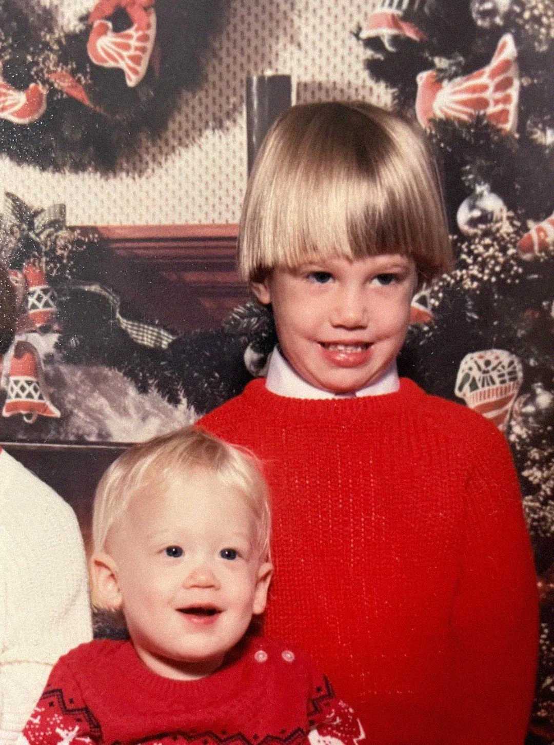 Two young children wearing red sweaters sit together in front of a festive holiday backdrop with wreaths and ornaments. The older child has a bowl haircut and smiles, while the younger child smiles with an open mouth.