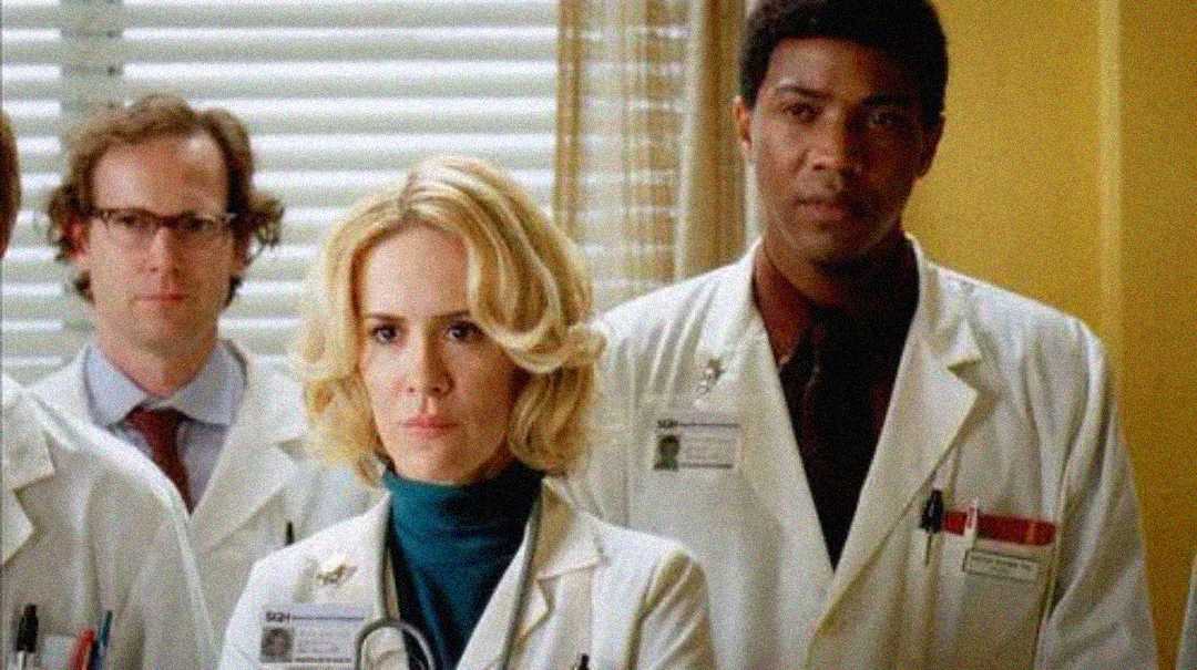 Three doctors wearing white lab coats and ID badges stand together in a hospital room, looking forward with serious expressions. Blinds cover the window behind them, and yellow walls are visible.