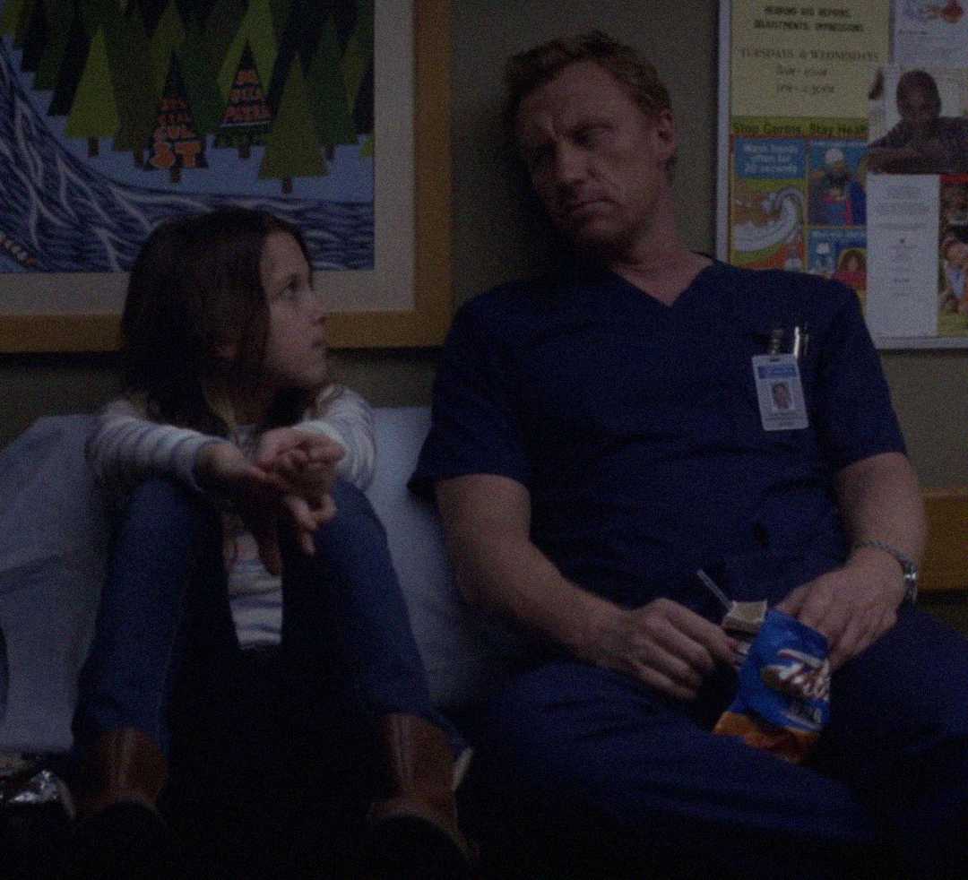 A young girl and a man in blue scrubs sit side by side on a bench in a dimly lit room, looking at each other. The man holds a snack bag, and children's artwork and posters are visible on the wall behind them.