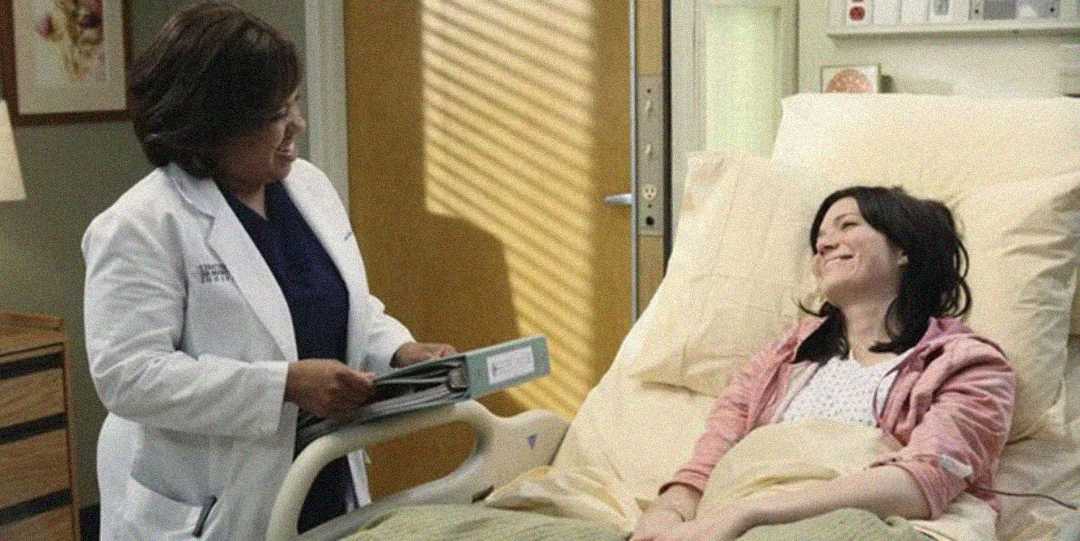 A doctor wearing a white coat stands by a hospital bed, smiling and holding a chart, while a female patient in a pink jacket lies in bed, also smiling, in a hospital room.