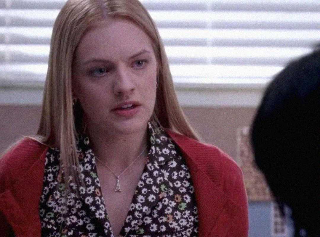 A young woman with long blonde hair, wearing a floral blouse and a red cardigan, sits indoors in front of a window with blinds, looking concerned or serious.