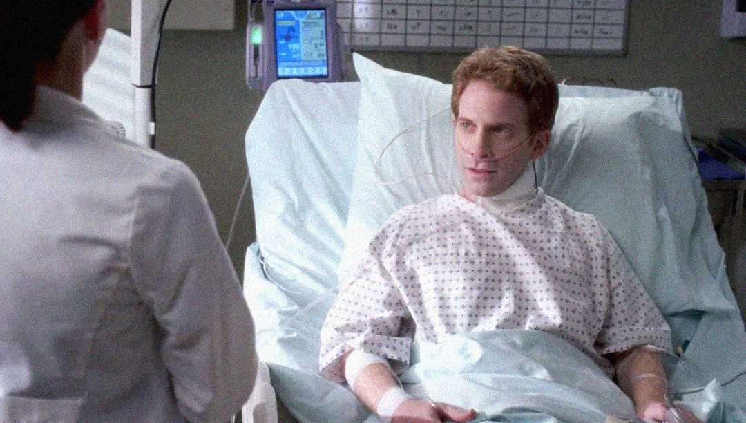 A man wearing a hospital gown lies in a hospital bed with an oxygen tube in his nose, looking at a person in a white coat who is standing nearby. Medical equipment and a chart are visible in the background.