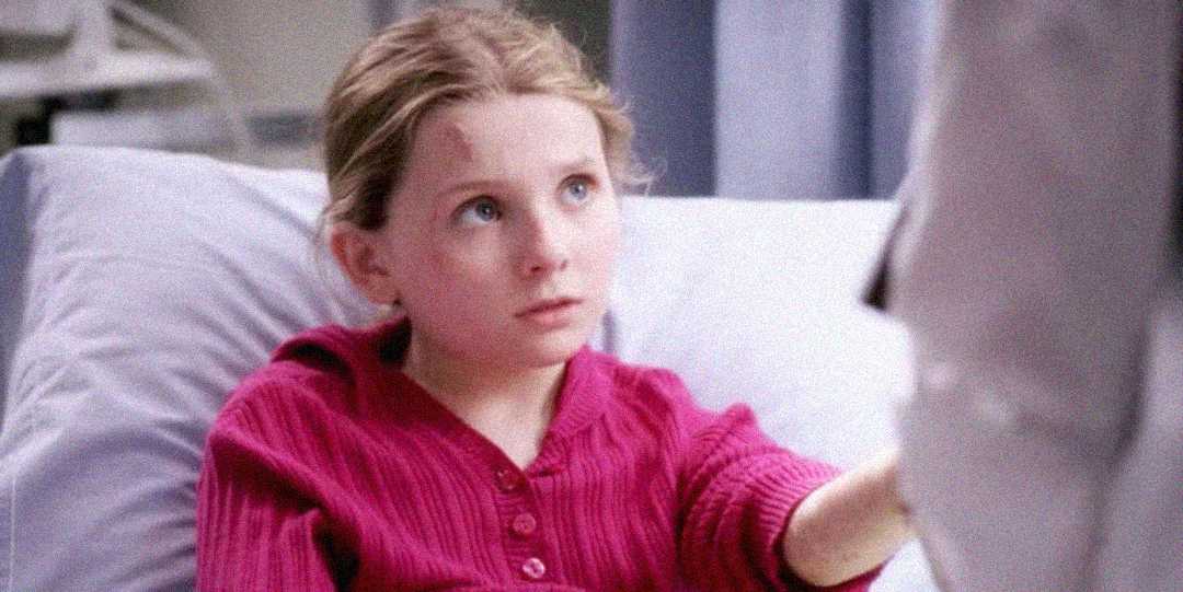 A young girl with blonde hair, wearing a pink sweater, sits up in a hospital bed looking up with a worried expression while holding the hand of someone next to her.