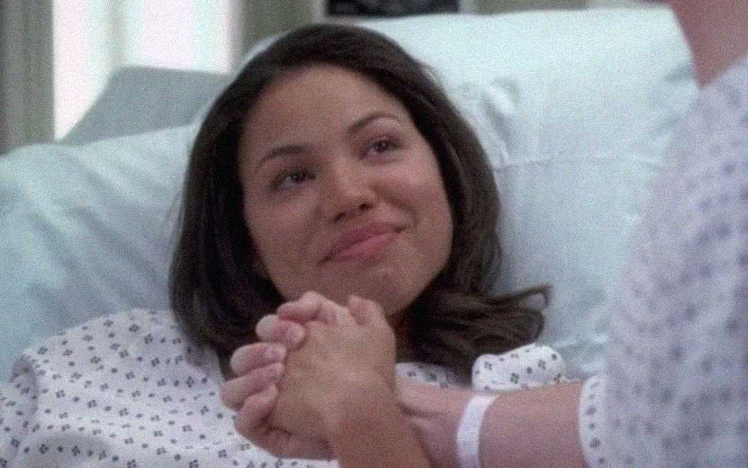 A woman in a hospital gown lies in a bed, smiling warmly as she holds hands with someone sitting beside her.