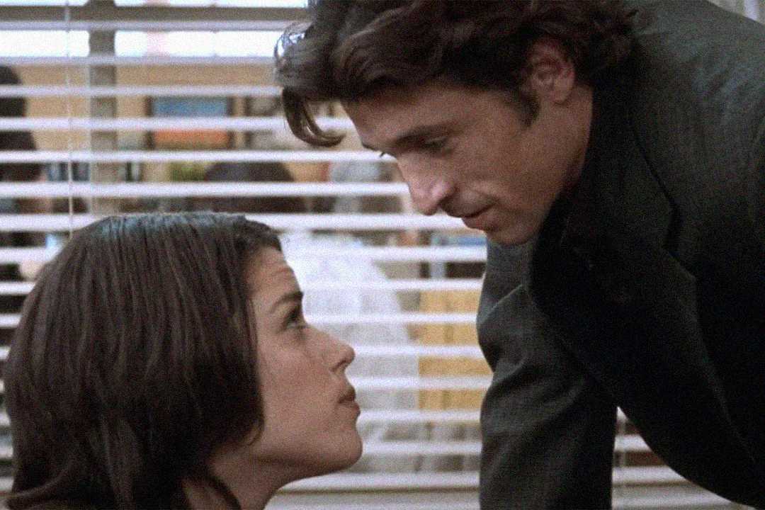 A woman with dark hair sits and looks up at a man with wavy hair who is leaning toward her. They are indoors with venetian blinds in the background, and both appear to be having a serious conversation.