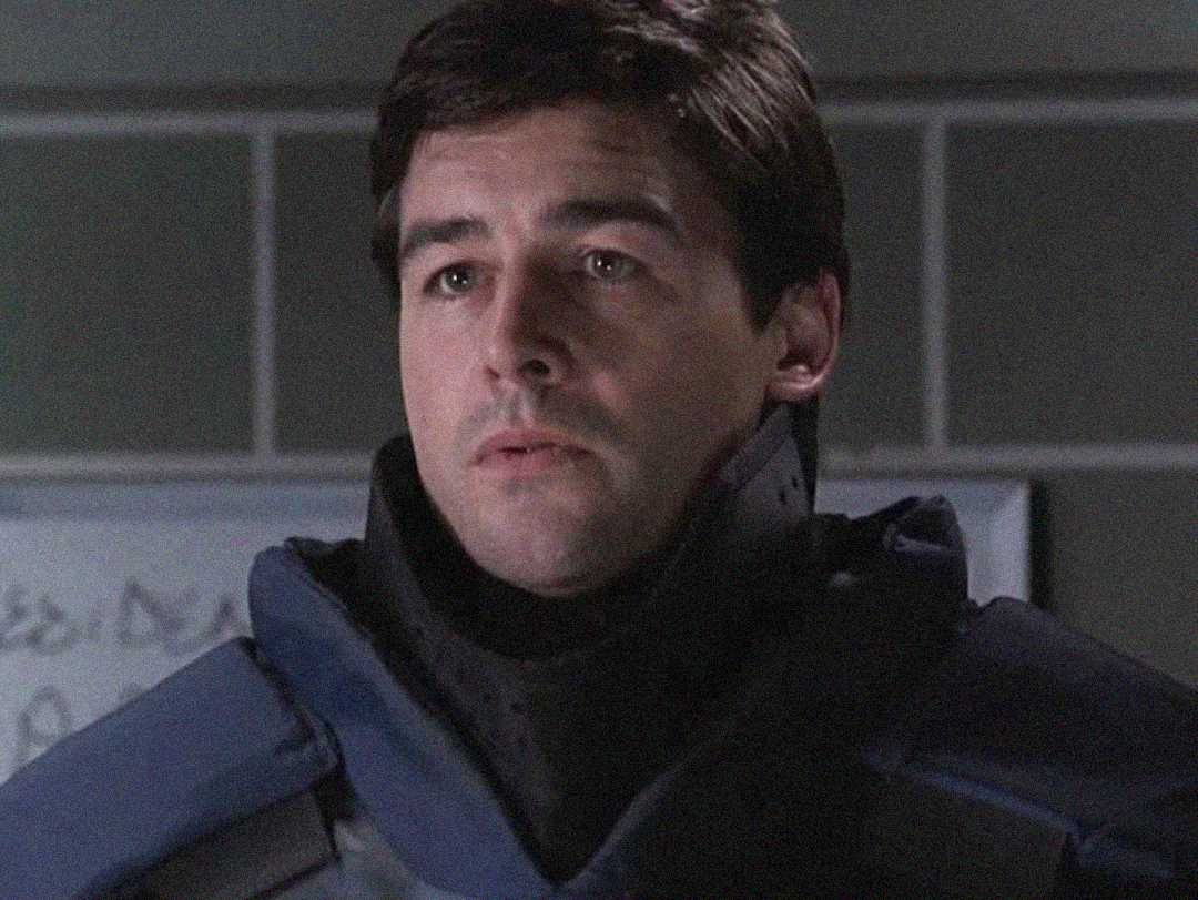 A man with dark hair wearing a heavy, padded bomb squad suit stands indoors in front of a tiled wall, looking slightly concerned.